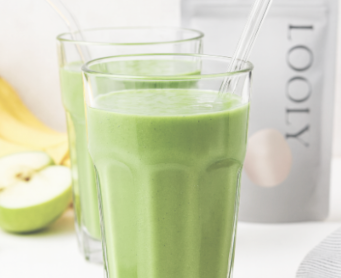 Zwei Gläser mit grünem Smoothie, im Hintergrund ein Standbodenbeutel mit der Aufschrift „LOOLY“ sowie angeschnittenes Obst.