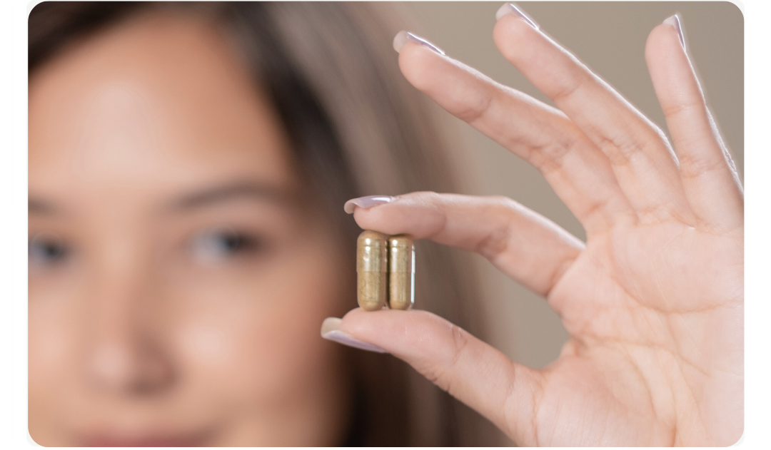 Person hält zwei Kapseln zwischen den Fingern, Gesicht im Hintergrund unscharf.
